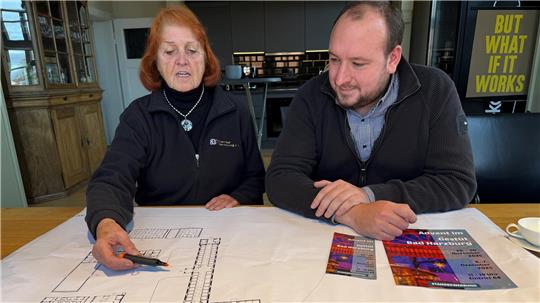 Zwei Personen sitzen an einem Tisch mit großen Bauplänen und Farbfächern, eine Person zeigt mit einem Stift auf die Pläne