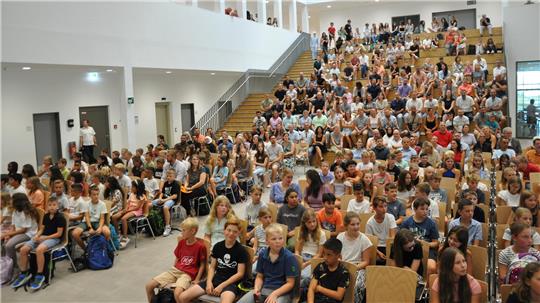 Große Gruppe von Kindern und Erwachsenen sitzt in einem modernen, hellen Auditorium mit Holztreppen und weißen Wänden