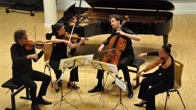 Vier Musiker sitzen im Halbkreis auf Stühlen und spielen Geige, Bratsche und Cello vor einem Flügel auf einer Bühne.