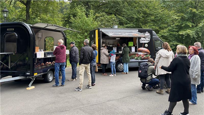 Menschen stehen in einer Schlange vor zwei Imbisswagen in einem Park mit Bäumen im Hintergrund.