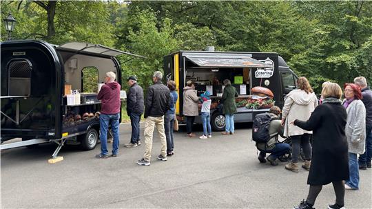 Menschen stehen in einer Schlange vor zwei Imbisswagen in einem Park mit Bäumen im Hintergrund.