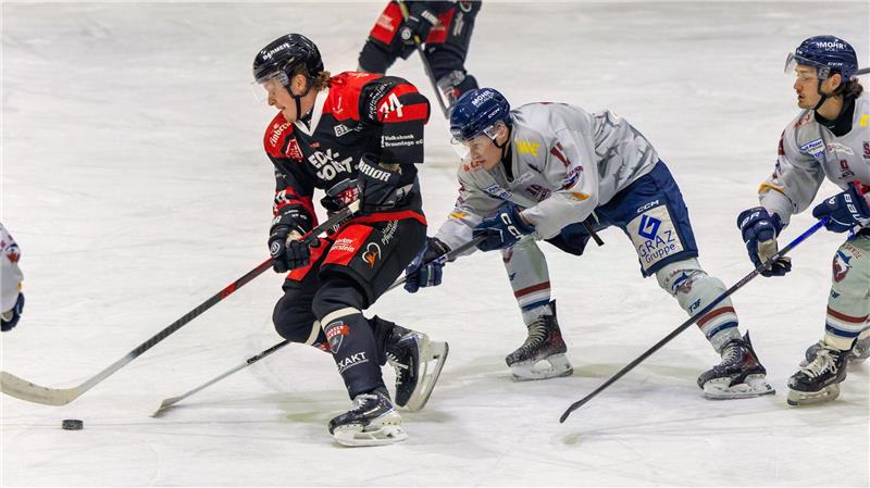 Eishockeyspieler in schwarzer und roter Ausrüstung mit Puck vor mehreren Gegenspielern in weißer und blauer Ausrüstung auf dem Eis.