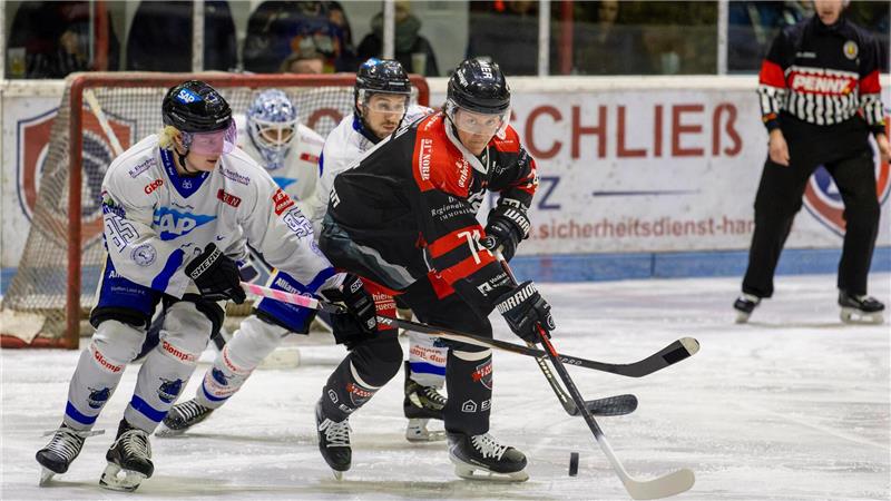 Drei Eishockeyspieler in Aktion auf dem Eis, zwei in weißen Trikots und einer in schwarzem Trikot, mit Schlägern und Puck vor dem Tor, im Hintergrund Schiedsrichter und Torwart