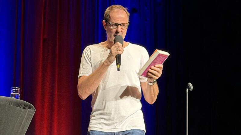 Mann mit Brille und weißem T-Shirt liest von einem Buch und spricht in ein Mikrofon auf einer Bühne mit rotem und blauem Hintergrund.