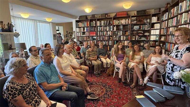 Das Bild zeigt viele Menschen in Stuhlreihen, eine Frau begrüßt sie, ringsherum sind viele Bücherregale. 