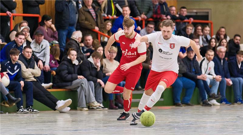 Zwei Hallenfußballspieler in rot-weißer Kleidung kämpfen um den Ball vor Zuschauern auf der Tribüne.