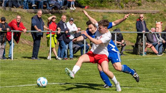 Zwei Fußballspieler in Aktion auf dem Spielfeld, einer in weißem Trikot und roter Hose, der andere in blauem Trikot, im Hintergrund Zuschauer an der Seitenlinie.