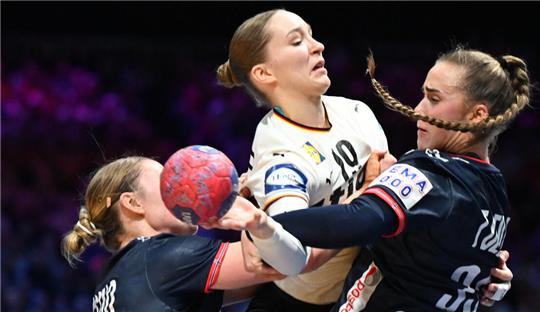 Drei Handballspielerinnen in Aktion, zwei in schwarzen Trikots verteidigen gegen eine Spielerin im weißen Trikot mit Ball.