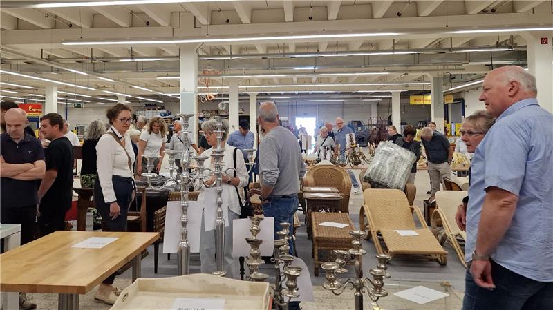 Mehrere Personen stehen in einer großen Halle mit Tischen, auf denen Kerzenhalter und andere Gegenstände ausgestellt sind.