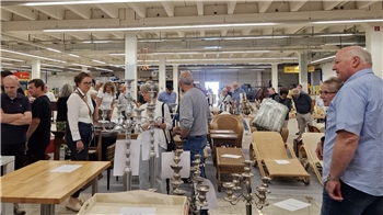 Mehrere Personen stehen in einer großen Halle mit Tischen, auf denen Kerzenhalter und andere Gegenstände ausgestellt sind.