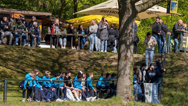 Gruppe von Kindern in blauen Jacken sitzt auf einer Wiese hinter einem Geländer, darüber stehen Erwachsene an einem Baum und unter einem gelben Sonnenschirm.