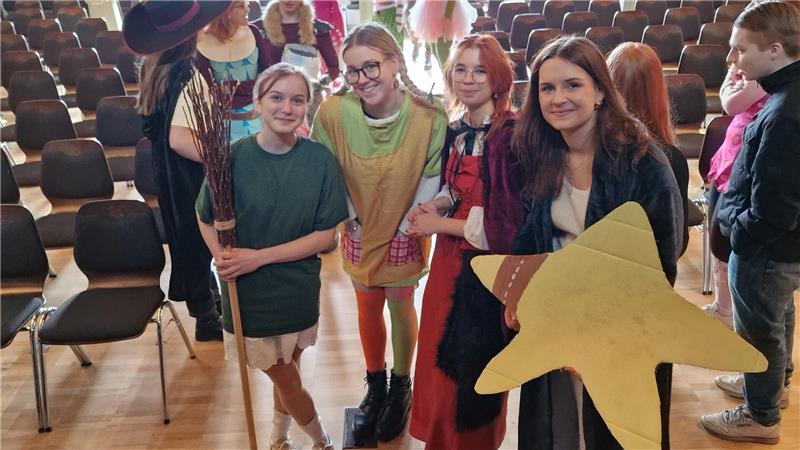 Gruppe von Jugendlichen in bunter Kleidung steht in einem Raum mit Reihen von Stühlen.