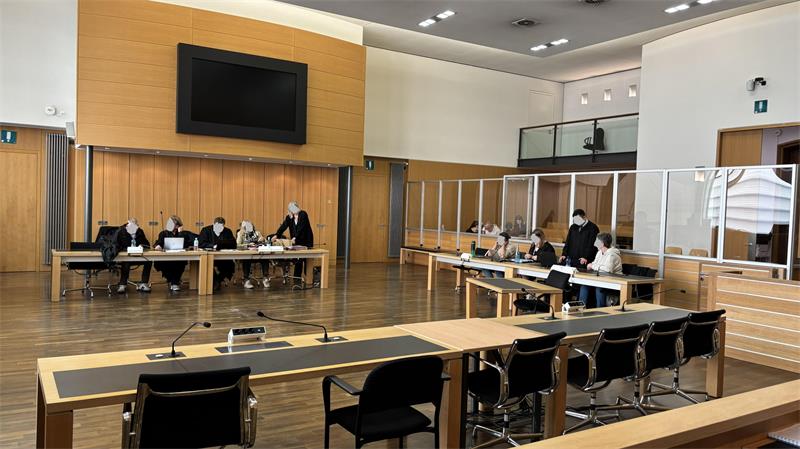 Großer Sitzungssaal mit mehreren Personen an Tischen, die durch transparente Trennwände getrennt sind, und einem großen Bildschirm an der Wand