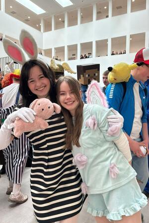 Zwei verkleidete Personen, eine mit gestreiftem Oberteil und Plüschbär, die andere mit Engelsflügeln und rosa Handschuhen, umgeben von weiteren kostümierten Menschen auf einer Veranstaltung.