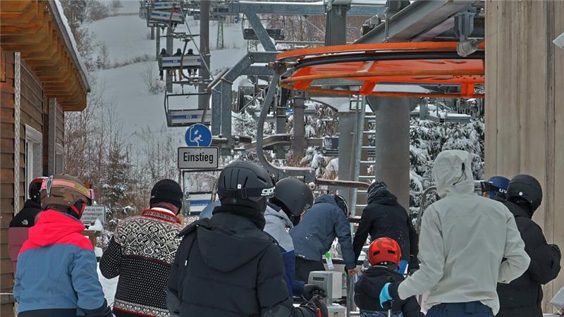 Mehrere Personen in Winterkleidung warten an einer Skiliftstation mit Schneelandschaft im Hintergrund.