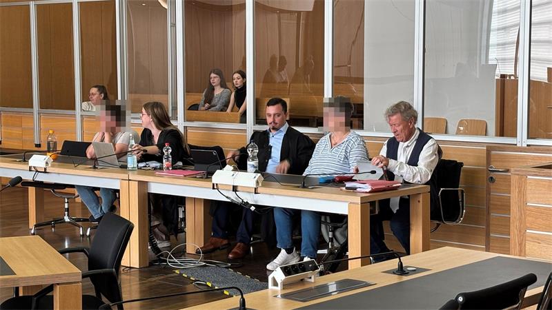 Fünf Personen sitzen an einem langen Tisch in einem Raum mit Holzvertäfelung und Glasabtrennung, zwei Personen sind verpixelt
