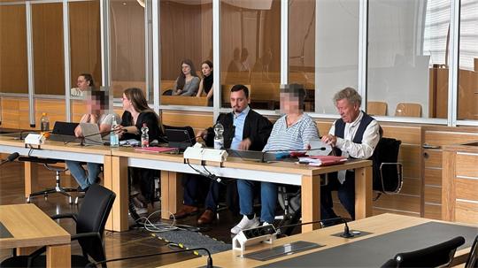 Fünf Personen sitzen an einem langen Tisch in einem Raum mit Holzvertäfelung und Glasabtrennung, zwei Personen sind verpixelt