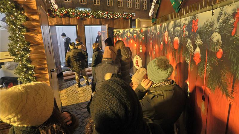 GZ-Redakteur Sowa im WC-Einsatz: Zwischen Klobürste und Flatrate Menschen in Winterkleidung stehen in einer Schlange vor einem festlich geschmückten WC-Holzstand mit Lichterketten und Tannenzweigen.