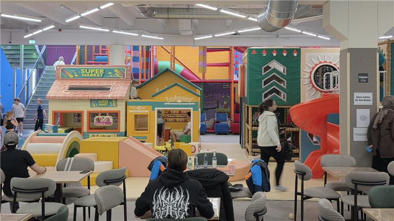 Innenraum eines Spielplatzes mit Klettergerüst, Rutsche, Spielhäusern und Sitzbereich mit Tischen und Stühlen, mehrere Personen sichtbar.