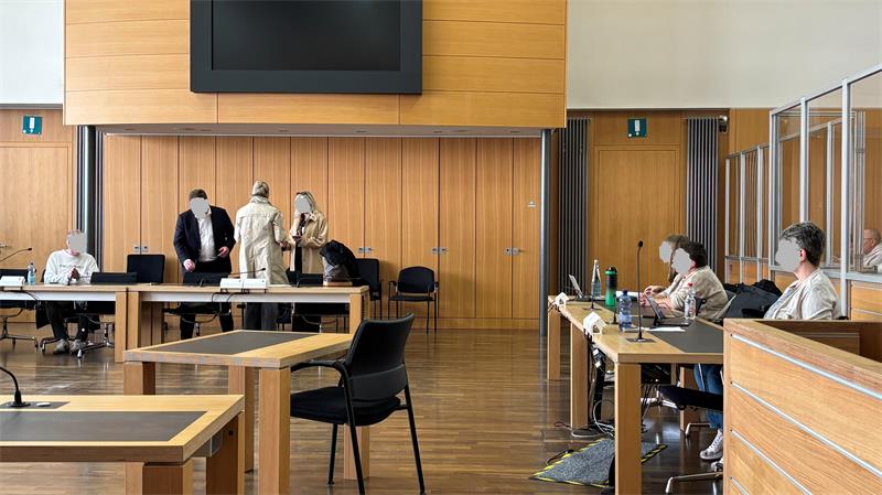 Mehrere Personen sitzen und stehen in einem großen Raum mit Holzwänden und Tischen, einige tragen Masken