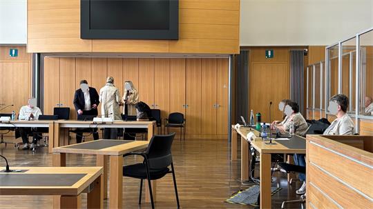 Mehrere Personen sitzen und stehen in einem großen Raum mit Holzwänden und Tischen, einige tragen Masken