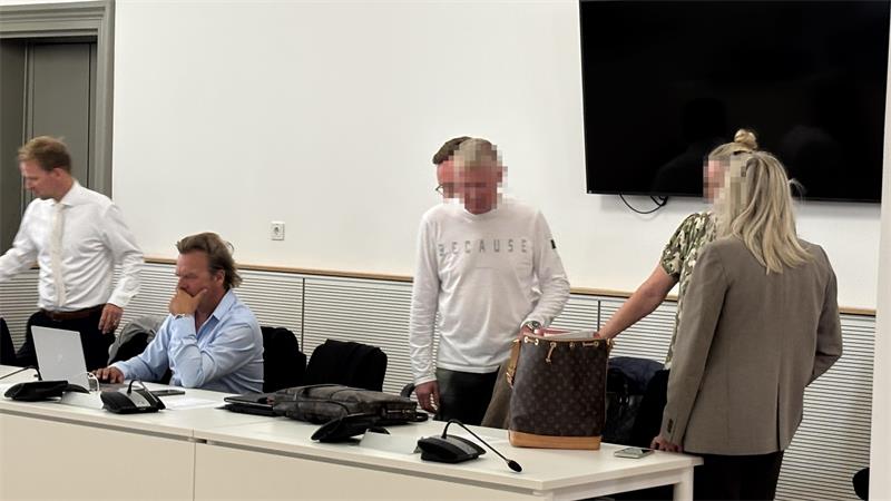 Fünf Personen in einem Besprechungsraum, drei sitzen an einem Tisch mit Laptops, zwei stehen, eine Person trägt eine weiße Sweatshirt mit Schriftzug