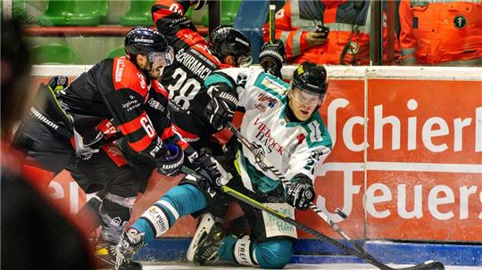 Eishockeyspieler in Aktion an der Bande, zwei in schwarzen Trikots und einer in weiß-grünem Trikot, alle mit Helm und Schläger auf dem Eis