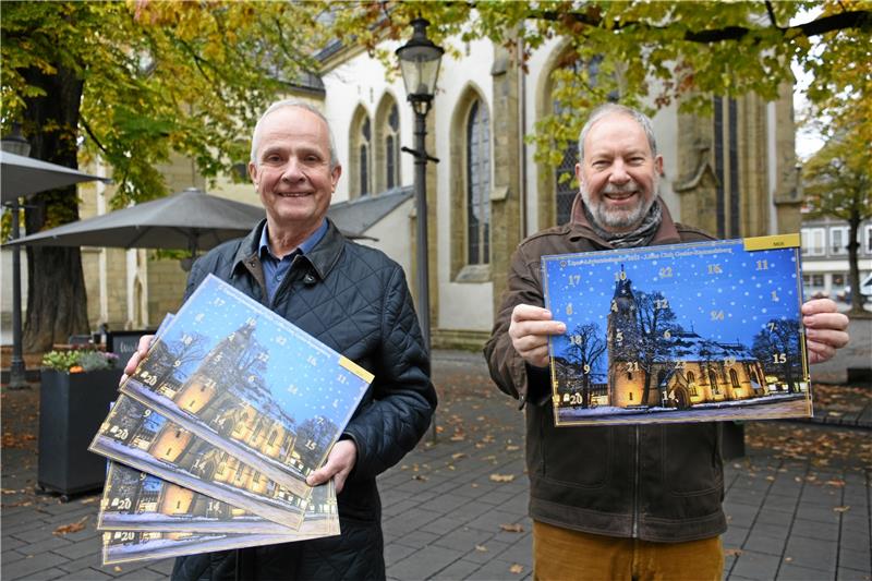 Lions-Adventskalender: Gehaltvolle Türchen vor der Jakobikirche Vor der St. Jakobikirche im Herbstlaub präsentieren (v.li.) Thomas Brych und Hartmut Prüssner vom Lions-Club Goslar-Rammelsberg den neuen Benefiz-Adventskalender 2023, der die heimelig beleuchtete Kirche im Winterkleid zeigt. Foto: Kempfer