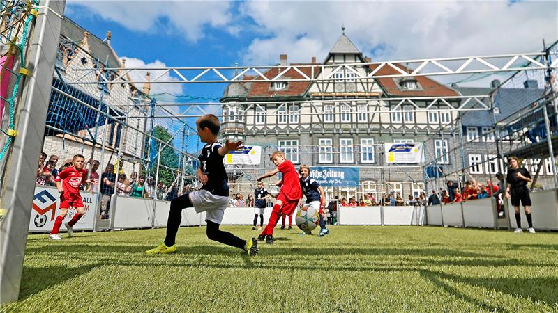 Kinder spielen Fußball auf einem kleinen Spielfeld vor einem historischen Fachwerkgebäude unter blauem Himmel