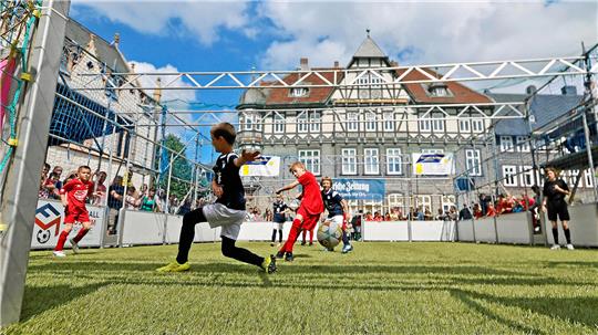 Kinder spielen Fußball auf einem kleinen Spielfeld vor einem historischen Fachwerkgebäude unter blauem Himmel