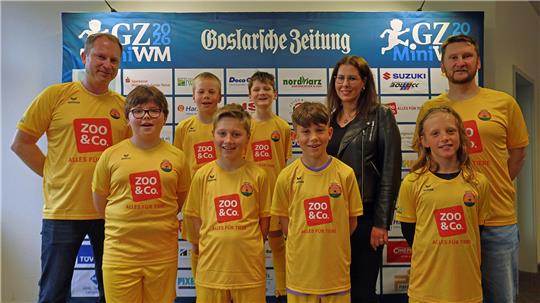 Gruppe von neun Personen, sechs Kinder in gelben Sporttrikots mit rotem "ZOO & Co."-Logo und eine Frau in schwarzer Jacke vor einer Werbewand mit "Goslarsche Zeitung"-Schriftzug