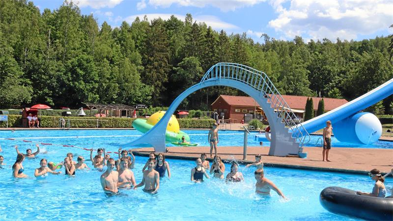 Menschen schwimmen und spielen in einem Freibad mit einer blauen Wasserrutsche und einem großen aufblasbaren Spielgerät im Hintergrund.