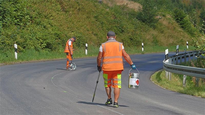 Zwei Personen in orangefarbener Warnkleidung markieren mit Kreide eine geschwungene Straße neben einer Leitplanke und Grünbewuchs.