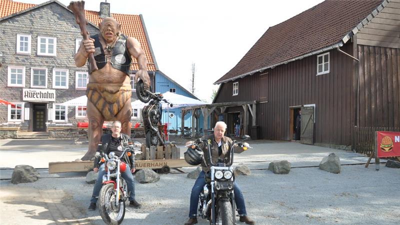 Zwei Männer auf Motorrädern vor einer großen Trollfigur und traditionellen Fachwerkhäusern