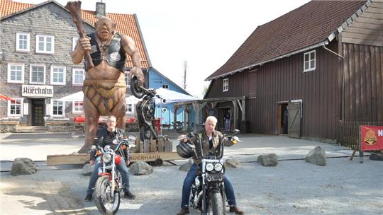 Zwei Männer auf Motorrädern vor einer großen Trollfigur und traditionellen Fachwerkhäusern