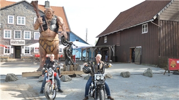 Zwei Männer auf Motorrädern vor einer großen Trollfigur und traditionellen Fachwerkhäusern