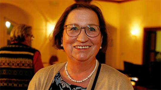 Lächelnde Frau mit Brille, Perlenkette und beigem Cardigan in einem warm beleuchteten Raum