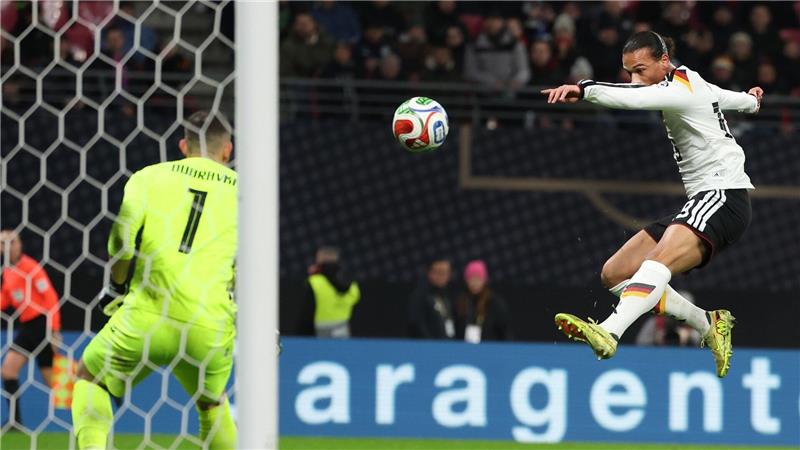 Der Nationalspieler Leroy Sané zielt im Sprung mit dem Ball aufs Tor, in dem ein Torhüter mit grünem Trikot steht.