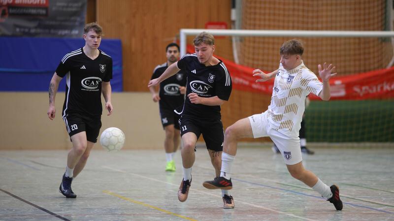 Drei Spieler in schwarzen Trikots und ein Spieler in weißem Trikot spielen Hallenfußball, der Spieler in Weiß führt einen Torschuss aus.