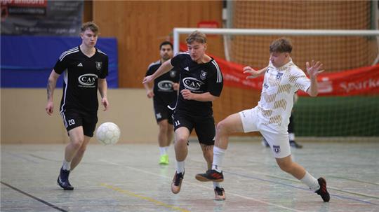 Drei Spieler in schwarzen Trikots und ein Spieler in weißem Trikot spielen Hallenfußball, der Spieler in Weiß führt einen Torschuss aus.
