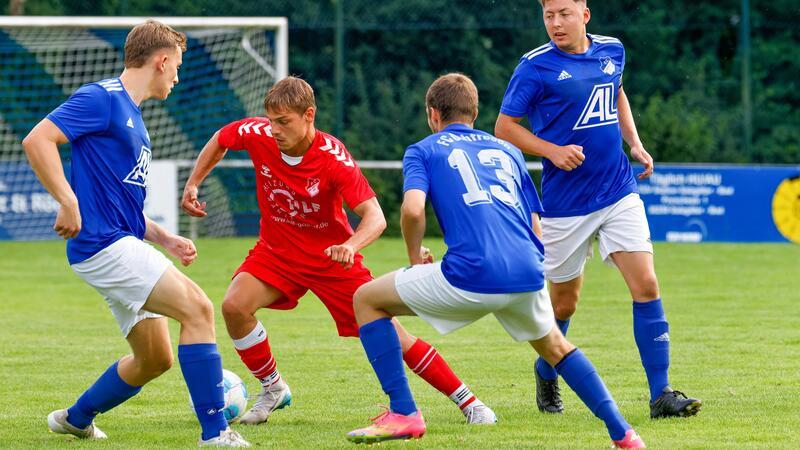 Vier Fußballspieler auf dem Spielfeld, zwei in roten Trikots und zwei in blauen Trikots, in Aktion vor einem Tor mit Netz im Hintergrund