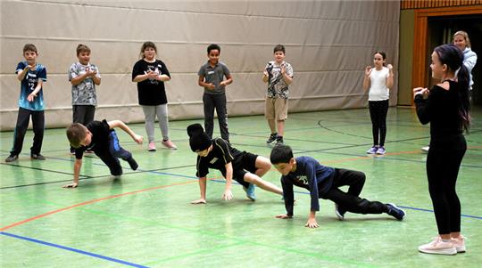 Drei Kinder führen Tanzschritte aus, während drumherumstehende Kinder sie anfeuern.