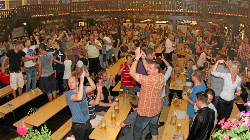 Menschen in einem Festzelt stehen an langen Holztischen und klatschen in die Hände.