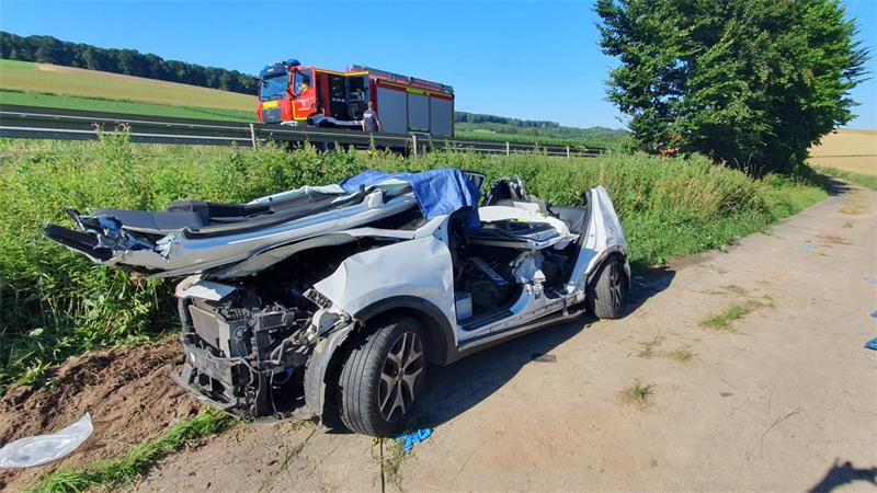 Schwer beschädigtes Auto ohne Türen steht auf einem Feldweg neben einer Straße mit einem Feuerwehrfahrzeug im Hintergrund