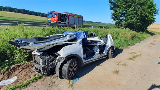 Schwer beschädigtes Auto ohne Türen steht auf einem Feldweg neben einer Straße mit einem Feuerwehrfahrzeug im Hintergrund