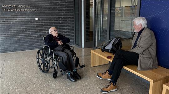 Person im Rollstuhl und sitzende Person auf einer Holzbank in einem Raum mit grauer Ziegelwand und blauer Wand.