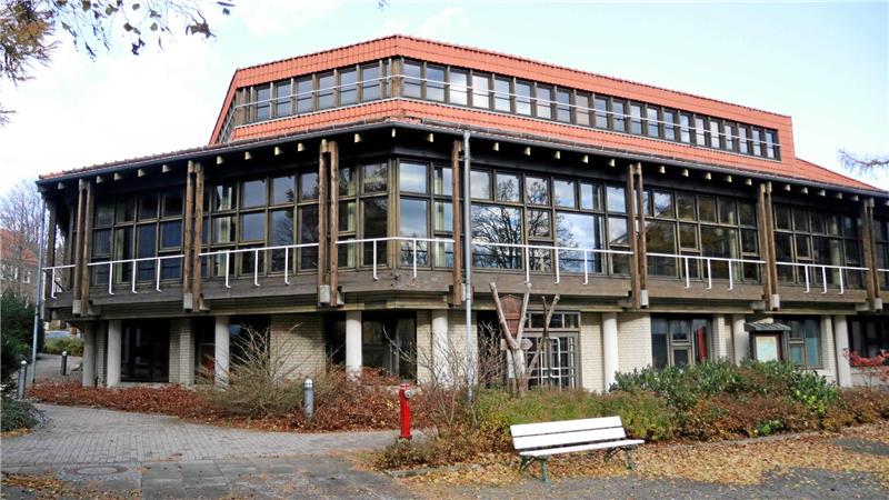Zweigeschossiges Gebäude mit großer Fensterfront, Holzverkleidung und rotem Ziegeldach, umgeben von herbstlicher Vegetation und einer weißen Parkbank im Vordergrund.