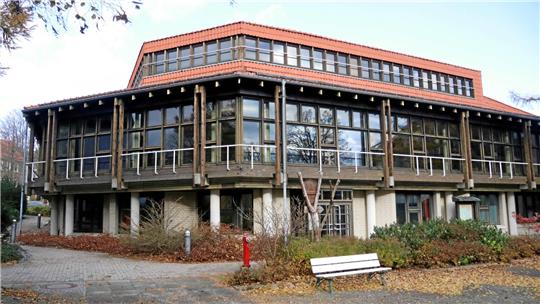 Zweigeschossiges Gebäude mit großer Fensterfront, Holzverkleidung und rotem Ziegeldach, umgeben von herbstlicher Vegetation und einer weißen Parkbank im Vordergrund.