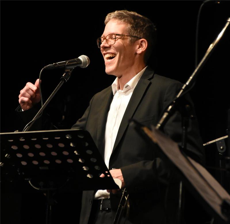 Ein vergnügter Mann lacht hinterm Notenständer am Mikrofon.