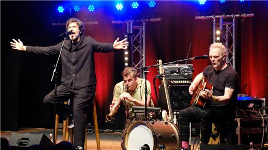 Drei Musiker auf Bühne mit rotem Hintergrund, einer singt mit ausgebreiteten Armen, einer spielt Trompete, einer Gitarre.